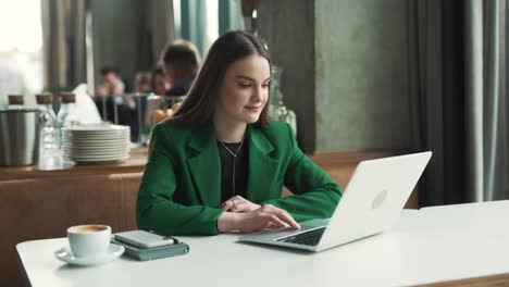 Eine-Hübsche-Frau-In-Einer-Grünen-Jacke-Beginnt-Zu-Tanzen,-Während-Sie-Gut-Gelaunt-Und-Lächelnd-An-Einem-Tisch-In-Einem-Restaurant-Sitzt