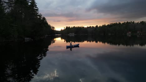 A-person-canoeing-on-a-still-lake-at-sunset,-with-the-sky's-vibrant-colors-reflecting-on-the-water,-surrounded-by-dense-trees