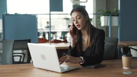 A-charming-woman-is-seated-in-the-office,-working-on-her-laptop-and-talking-on-the-phone