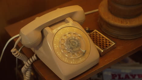 Altes-Weißes-Telefon-Im-Wählscheibenstil-Auf-Beistelltisch-Neben-Gebogener-Metallflasche-Bei-Holzlampe
