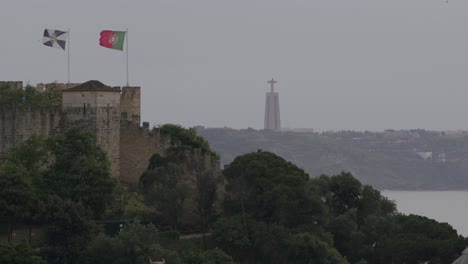 Toma-Estática-De-La-Bandera-De-Portugal-Ondeando-En-Un-Día-Ventoso-Con-Una-Estatua-De-Jesús-Al-Fondo-En-Lisboa,-Portugal.