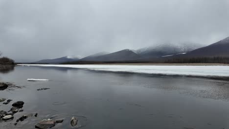 Slow-flow-of-another-half-of-the-frozen-river-against-the-background-of-a-gray-sky-and-snowy-mountains