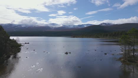 Aerial-flying-over-waters-of-Loch-Morlich-revealing-Cairngorms-mountains,-Scotland