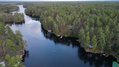 Aerial-view-of-a-person-canoeing-on-a-calm-forest-river,-surrounded-by-dense-trees-and-cabins-along-the-banks