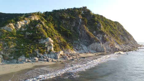 Santa-marta-beach-with-waves-lapping-the-shore,-lush-green-hillside,-sunny-day,-aerial-view