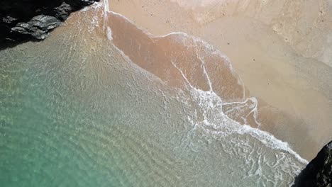 Cornish-Cove-in-Slow-Motion-with-Turquoise-Waves-Rolling-Over-a-Beach,-Aerial-Ascending-Drone,-UK