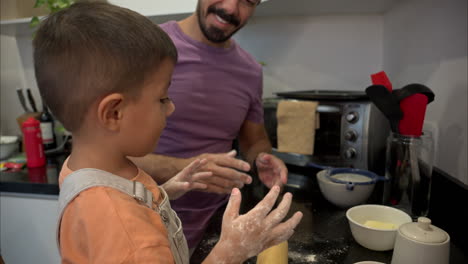 Mexican-latin-father-cleaning-his-son's-nose-covered-in-flour-while-they-prepare-a-cake-in-the-kitchen-having-fun