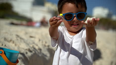 Zeitlupe-Eines-Coolen-Mexikanischen-Lateinamerikanischen-Kleinen-Jungen-Mit-Sonnenbrille-Und-Weißem-T-Shirt,-Der-In-Die-Kamera-Schaut-Und-Einige-Muscheln-Zeigt,-Die-Er-Am-Strand-In-Cancun-Gefunden-Hat