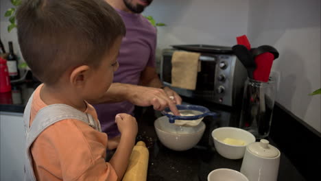 Junger-Mexikanischer-Lateinamerikanischer-Junge-Hilft-Seinem-Vater-In-Der-Küche,-Während-Sie-Mit-Einem-Sieb-Für-Das-Mehl-Kekse-Backen