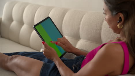 Mexican-latin-girl-with-a-pink-top-laying-on-her-sofa-browsing-on-her-tablet-using-her-index-finger-from-right-to-left