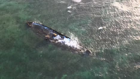 Drone-aerial-moving-around-a-shipwreck-in-the-ocean-with-waves-crashing-into-it-during-sunset