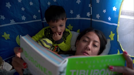 Close-up-of-a-mexican-latin-brunette-mother-reading-a-book-to-her-son-laying-inside-his-castle-tent-having-a-moment-together