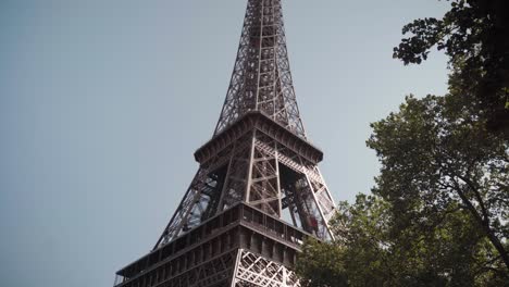 Una-Toma-De-ángulo-Bajo-De-La-Torre-Eiffel-Se-Eleva-Majestuosamente-Contra-Un-Cielo-Despejado-De-Verano,-Enmarcada-Por-Exuberantes-árboles-Verdes-En-Primer-Plano