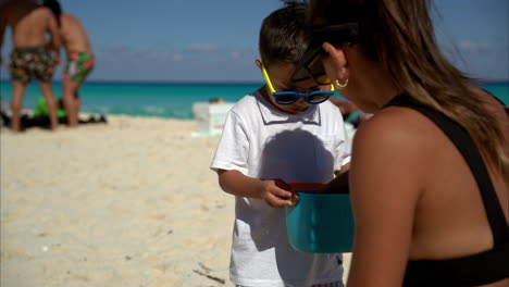 Süßer-Mexikanischer-Lateinamerikanischer-Brünetter-Junge-Mit-Sonnenbrille-Und-Weißem-T-Shirt-Auf-Der-Suche-Nach-Muscheln-Mit-Seiner-Mutter-An-Einem-Strand-In-Cancun,-Mexiko