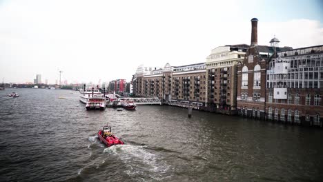 A-vibrant-red-boat-navigates-a-river-in-front-of-historic-buildings-in-London,-showcasing-a-lively-waterside-cityscape