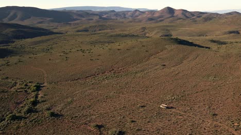 Drone-shot-of-a-safari-vehicle-driving-over-that-vast-and-arid-landscape-of-the-Klein-Karoo-in-South-Africa,-with-the-Lanberg-mountains-in-the-distance