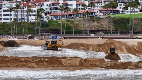 Bulldozer-Arbeiten-An-Der-Sandverlagerung-An-Einem-Strand-Mit-Küstenhäusern-Und-Palmen-Im-Hintergrund