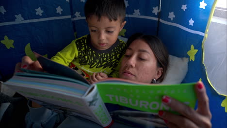 Close-up-of-a-young-mexican-latin-boy-and-his-brunette-mother-reading-a-book-laying-inside-his-castle-tent-having-a-moment-together