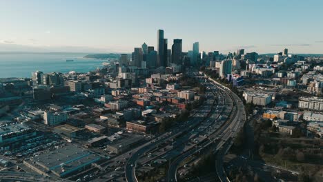 High-Drone-Flyover-of-Afternoon-Traffic-with-Long-Shadows,-Seattle-Skyline,-and-Puget-Sound-Backdrop