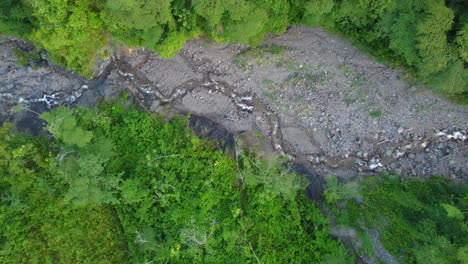 A-top-down-birds-eye-view-following-a-dry-riverbed-in-a-rainforest-with-a-small-stream-of-flowing-water,-Risaralda,-Colombia