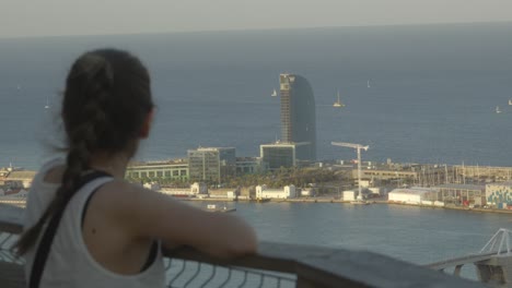 Mujer-Caucásica-Parada-En-Un-Balcón-Con-Vistas-Al-Puerto-De-Barcelona-Con-Un-Hotel-Icónico