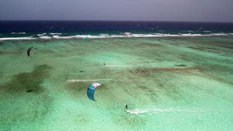 Kite-surfers-gliding-over-clear-turquoise-waters-by-a-barrier-reef,-vibrant-and-active-scene,-sunny-day,-aerial-view