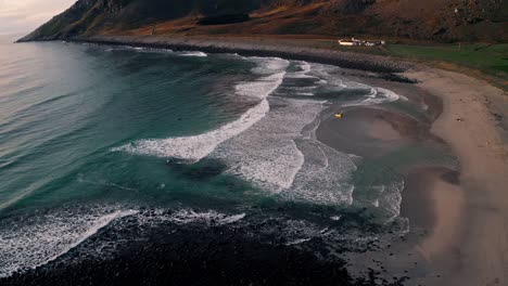 Aerial-view,-orbit-pan,-of-the-waves-crashing-at-Unstad-Beach,-while-surfers-enjoy-the-gorgeous-sunset,-on-a-clear-blue-sky-day,-Lofoten-Islands,-Norway