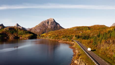 Flying-through-the-golden-leaves-of-trees-and-above-a-lake,-orbiting-around-a-white-camper-van-making-its-way-on-the-road,-Lofoten-Islands,-Norway