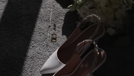 White-high-heeled-shoes,-wedding-rings,-and-necklace-on-a-carpeted-floor-in-sunlight