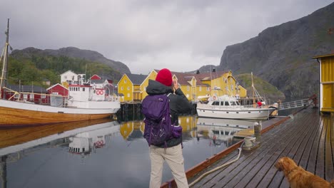 SLOW-MOTION-Push-out-reveal-of-a-young-female-photographer-tourist-and-her-golden-retriever-dog-photographing-the-beautiful-fisher-village-of-Å-and-its-red-rorbu-houses-in-the-Lofoten-Islands,-Norway