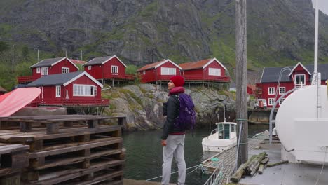 SLOW-MOTION-Push-out-shot-of-a-young-female-photographer-tourist-admiring-the-beautiful-fisher-village-of-Å-and-its-red-rorbu-houses-in-the-Lofoten-Islands,-Norway