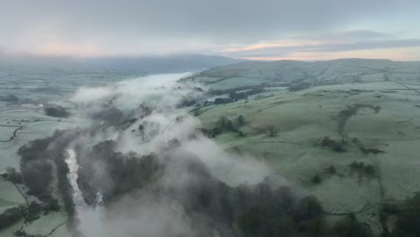 Misted-patchwork-field-hillside-with-river-shrouded-by-mist-and-dawn-glow-on-the-horizon-at-sunrise-in-winter