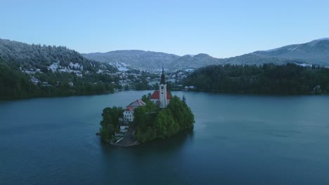 Luftaufnahme-Einer-Kleinen-Grünen-Insel-Mit-Der-Kirche-Mariä-Himmelfahrt-Inmitten-Des-Ruhigen-Bleder-Sees,-Slowenien