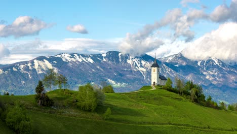 Picturesque-scene-of-a-solitary-church-perched-on-a-verdant-hill,-framed-by-the-grandeur-of-snow-capped-mountains-and-a-sky-streaked-with-fast-moving-clouds-during-sunny-day