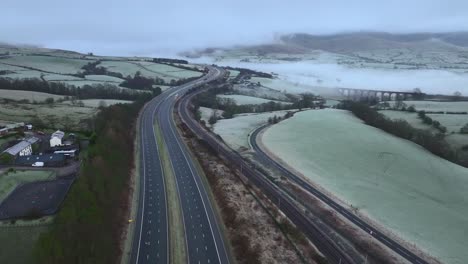 Volando-Sobre-El-Puente-En-La-Tranquila-Autopista-M6-Hacia-Colinas-Cubiertas-De-Niebla-Al-Amanecer-En-Invierno-Con-Un-Viaducto-De-Piedra-En-La-Distancia