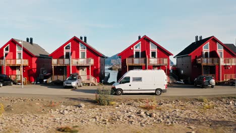 Seitliche-Kamerafahrt-Eines-Weißen-Wohnmobils-Vor-Wunderschönen-Roten-Rorbu-Häusern,-Die-Die-Beeindruckende-Bergkulisse-Von-Henningsvær,-Lofoten-Inseln,-Norwegen-Zeigt