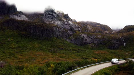 Aerial-footage-of-a-white-campervan-with-gorgeous-mountains-in-the-background,-leaving-the-beautiful-fisher-village-of-Å,-in-the-Lofoten-Islands,-Norway