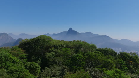 Close-aerial-footage-flying-over-the-trees-of-a-mountain-with-Botafogo-neighborhood-in-Christ-Redeemer-statue-in-the-background-in-Rio-de-Janeiro