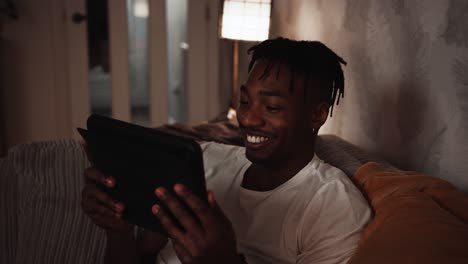 Young-black-male-watches-and-smiles-at-his-tablet-device-while-sat-on-the-sofa