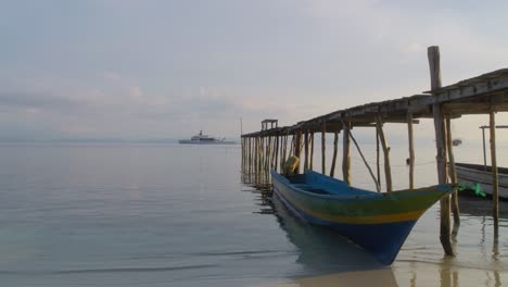 Kleines-Holzboot-Vor-Anker-In-Raja-Ampat-Am-Steg-Mit-Luxus-Superyacht-Wanderlust-Im-Hintergrund