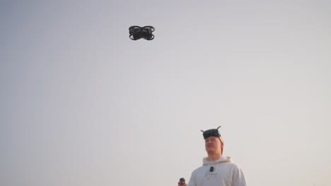 Low-angle-view-of-drone-hover-in-air-and-guy-with-FPV-goggles,-empty-sky-behind