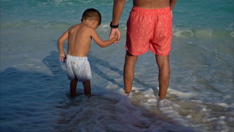 Slow-motion-of-a-mexican-latin-father-and-his-young-son-holding-hands-standing-on-the-sand-being-hit-by-a-wave-in-Cancun