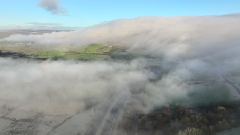 Flying-above-low-drifting-cloud-and-mist-with-fog-moving-down-background-hillside-near-M6-motorway-at-dawn-in-winter