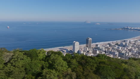 A-fast-aerial-fly-over-the-trees-of-a-mountain-top,-rotating-to-reveal-the-buildings-and-beach-of-Copacabana-in-Rio-de-Janeiro