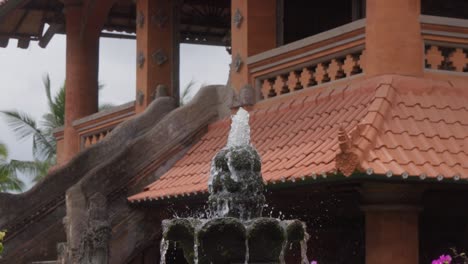 Traditional-stone-fountain-with-cascading-water,-surrounded-by-vibrant-flowers,-stands-in-front-of-traditional-Balinese-architecture