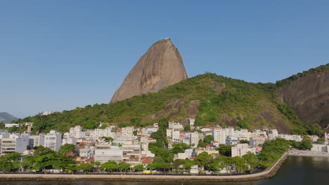 Aerial-footage-flying-a-backwards-over-the-water,-revealing-a-neighborhood-with-cars-driving-on-a-road-with-Sugarloaf-mountain-in-the-background-in-Rio-de-Janeiro-Brazil