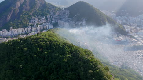 Aerial-footage-flying-over-the-trees-of-mountaintop-with-smoke-and-favelas-in-the-distance-in-Rio-de-Janeiro-Brazil