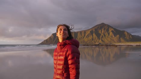 SLOW-MOTION,-a-beautiful-young-woman-admiring-the-view-and-smiling-on-the-Flakstad-beach-in-front-of-a-gorgeous-golden-sunset,-Lofoten-Islands,-Norway