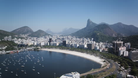 Ariel-Zeitraffer-Des-Strandes-Der-Botafogo-Bucht-Mit-Segelbooten-Und-Verkehr,-Der-Tagsüber-Herumfährt,-Mit-Der-Christusstatue-Im-Hintergrund-In-Rio-De-Janeiro