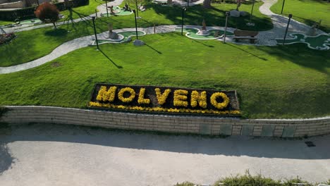 Ascending-aerial-looks-down-on-town-name-written-in-flowers,-Molveno,-Italy